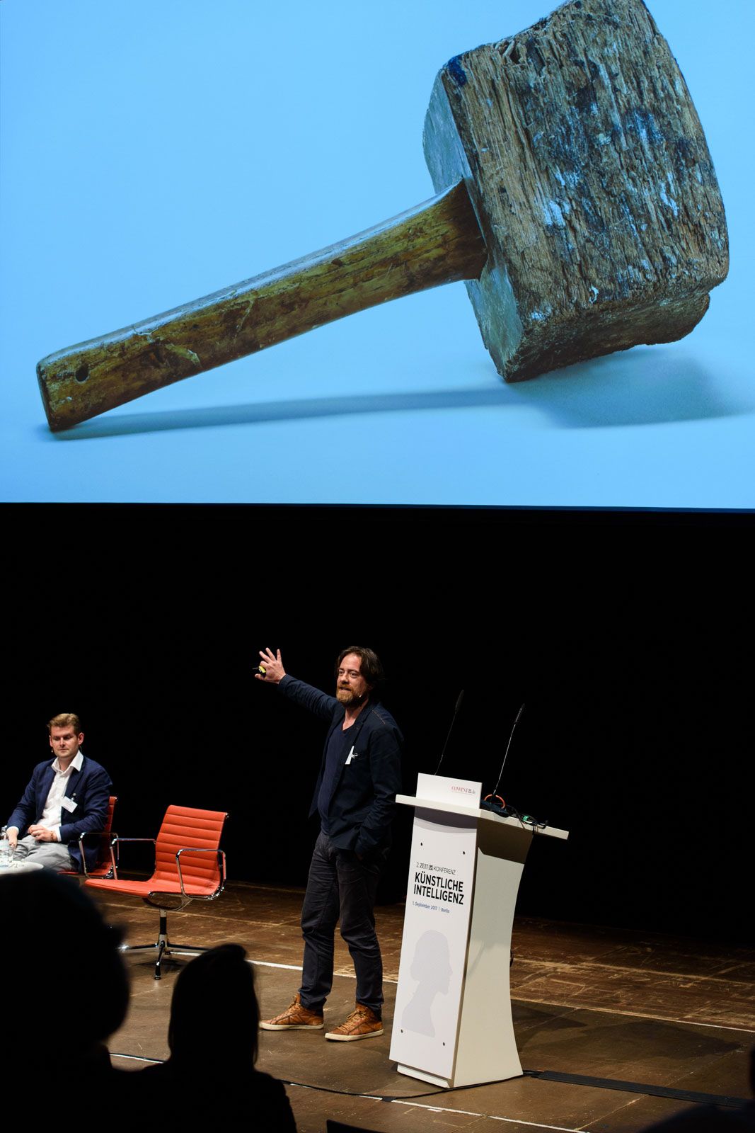 Philipp Schäfer steht gestikulierend am Rednerpult einer Konferenzbühne; hinter ihm füllt die Projektion eines alten hölzernen Schlegels die Bühnenwand, am Pult steht „Künstliche Intelligenz“.